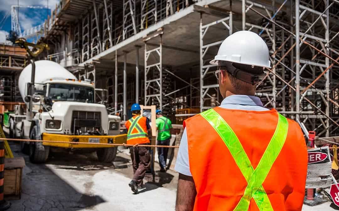 Man in orange reflector vest on construction site overseeing two men working